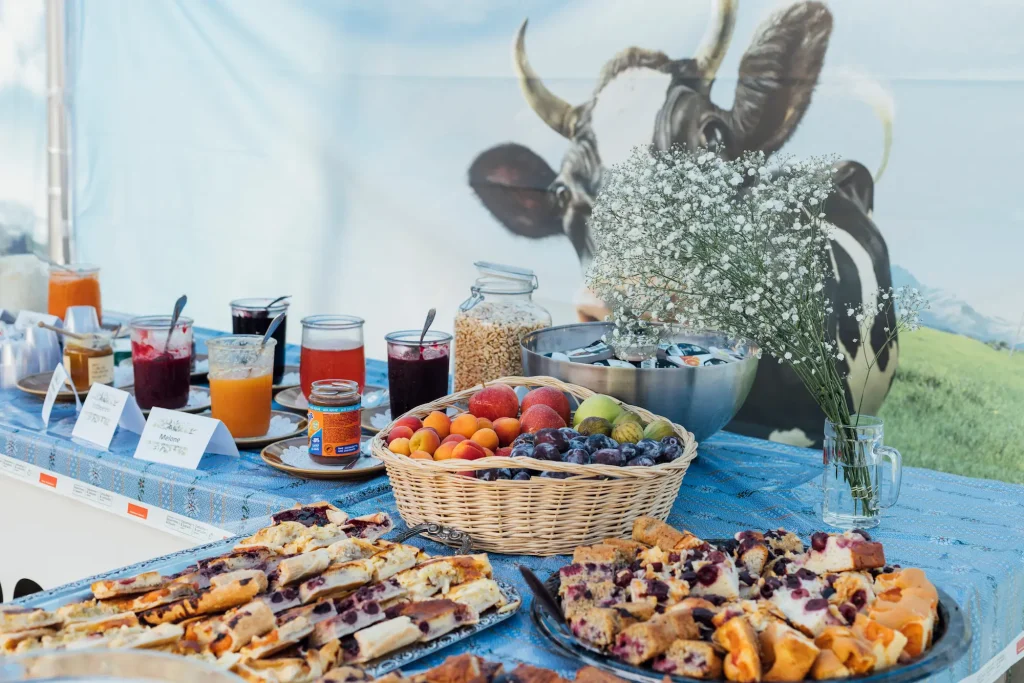 Buffet per la colazione con marmellate, succhi di frutta e croissant, con un poster raffigurante una mucca sullo sfondo.